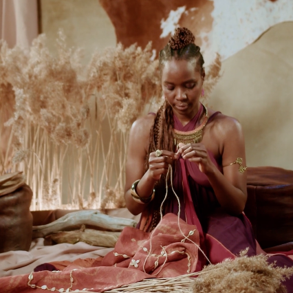 A woman with braided hair sits on the floor surrounded by fabrics and holding thread.